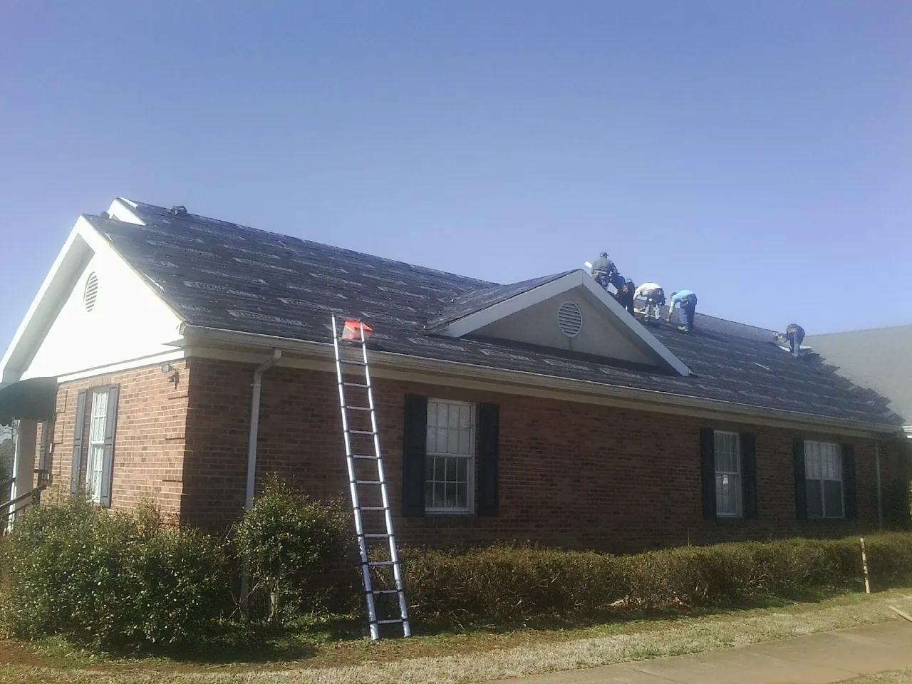 Crew tearing shingles off a brick ranch roof with a ladder set — Emergency Roof Repair work by our crew in Latimer